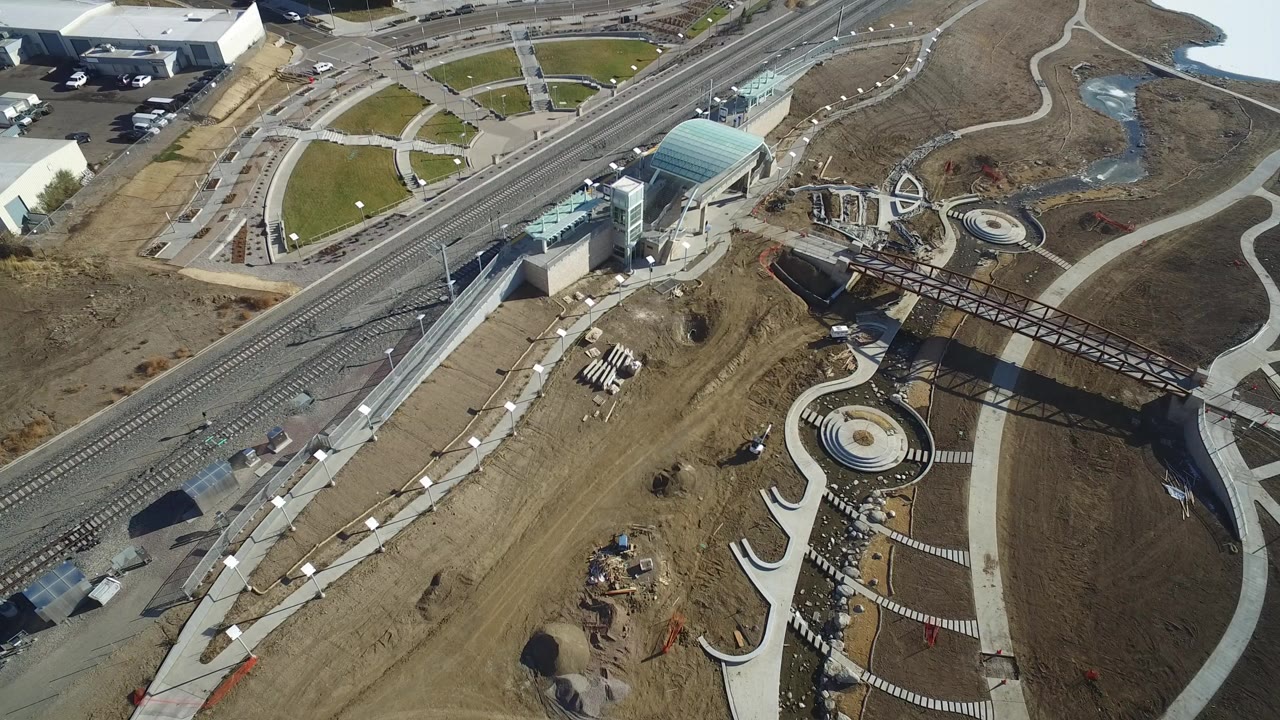 Opening overhead frame in Hidden Lake, Colorado showing diagonal rail tracks, a glass-roofed station, curved pedestrian paths, a circular plaza, bare earth, and no visible people.