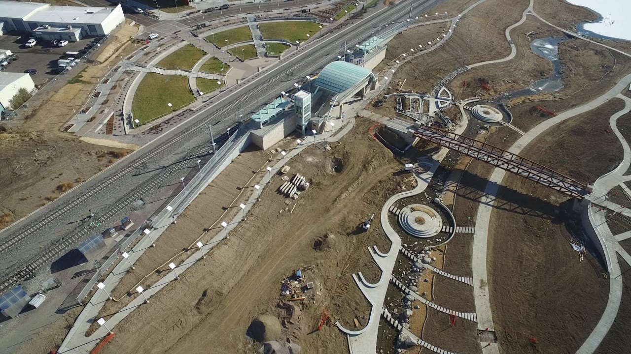 Drone view over tracks, station buildings, a pedestrian bridge, paths, and open land.