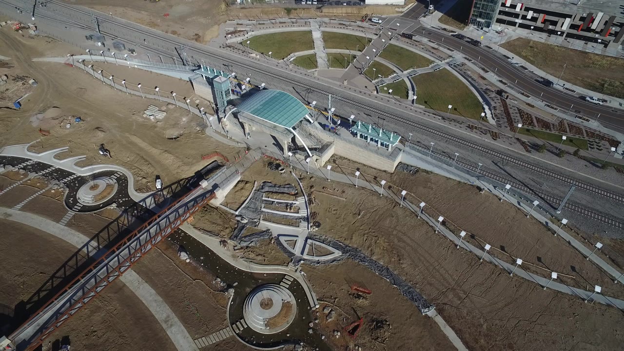 Overhead view in Hidden Lake, Colorado showing rail tracks beside a teal-roofed pavilion, circular paved areas, construction dirt, and a rust-colored pedestrian bridge.