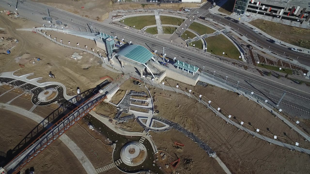 High aerial frame in Hidden Lake, Colorado with the teal-roofed structure centered near rail lines, curved walkways, bare construction ground, and roads at the top of frame.