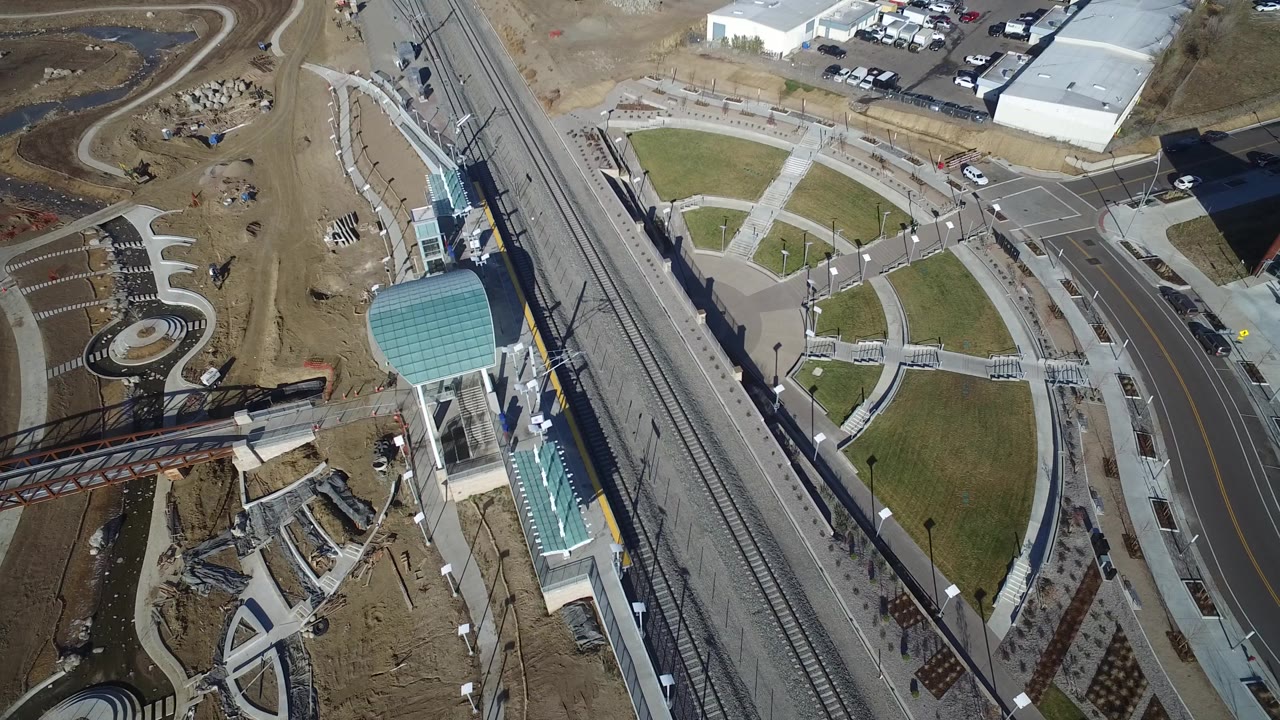 A high overhead view in Hidden Lake, Colorado shows the rail corridor running beside a curved roadway, circular plaza paving, brown landscaping, and nearby industrial lots with no visible people.