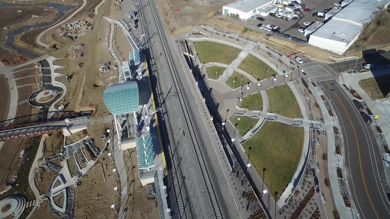 The aerial view over Hidden Lake, Colorado places rail tracks and a roadway beside a green lawn, paved paths, parking areas, and white commercial buildings, with no visible people.