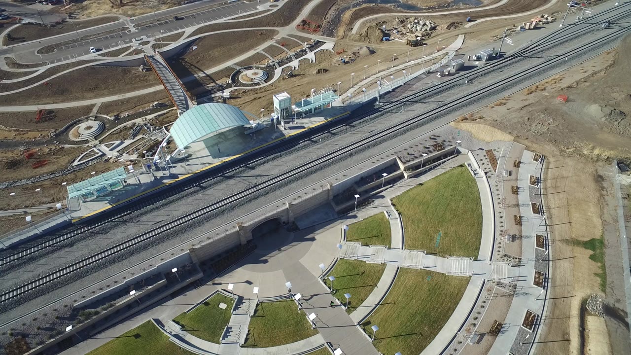 Opening aerial frame over Hidden Lake, Colorado showing rail tracks crossing the scene, a pale domed structure near the upper left, curved paths, green lawn panels, bare ground, and no visible people.