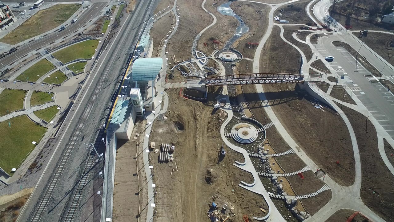 Aerial view of rail tracks, a glass-roofed structure, roadways, dirt construction areas, and curving concrete paths.