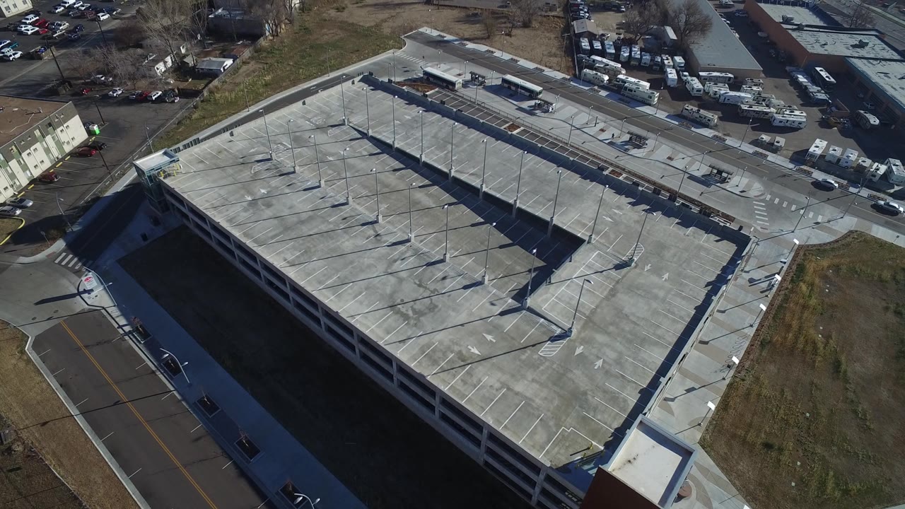 Aerial frame over Hidden Lake, Colorado, with the empty parking deck centered, rooftop-like concrete surfaces, light poles casting shadows, industrial lots at the top edge, and no visible people.