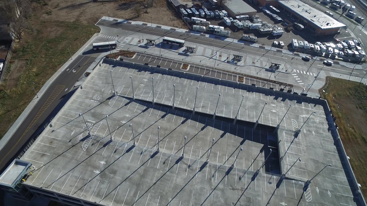 High aerial frame over a concrete parking garage in Hidden Lake, Colorado, with painted stalls, light poles, roads, buses, parked vehicles, and no visible people.