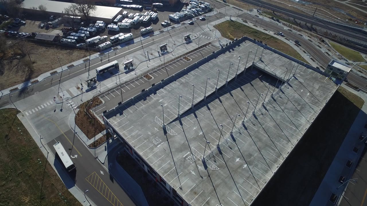 Oblique aerial frame over a large concrete parking deck in Hidden Lake, Colorado, with buses near the road, rail tracks beyond, winter-brown grass, and no visible people.