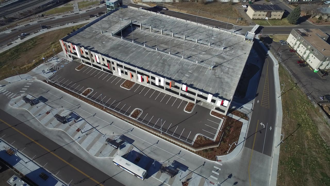 The aerial view continues across the large building in Hidden Lake, Colorado, with the parking lot in front, roof framing above, a road along the side, distant transport lines, and no visible people.