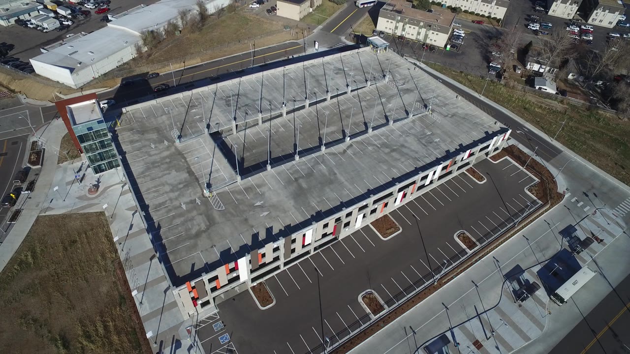 Oblique aerial frame of the Hidden Lake shopping center, showing more of the roof deck lanes, tall light poles, front facade, paved parking rows, and no visible people.