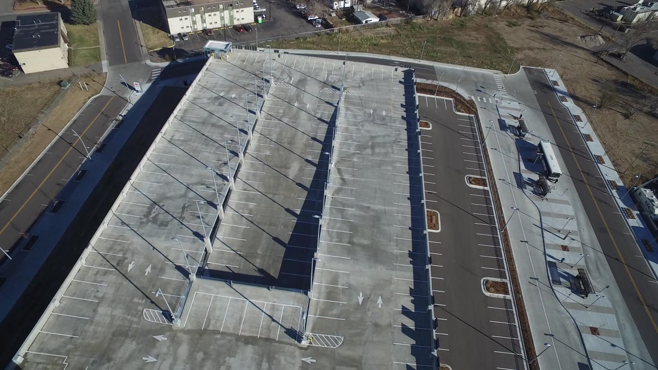 High aerial frame in Hidden Lake, Colorado, with the gray work-site roof or deck centered, parking bays along both sides, an adjacent street, small buildings, and no visible people.