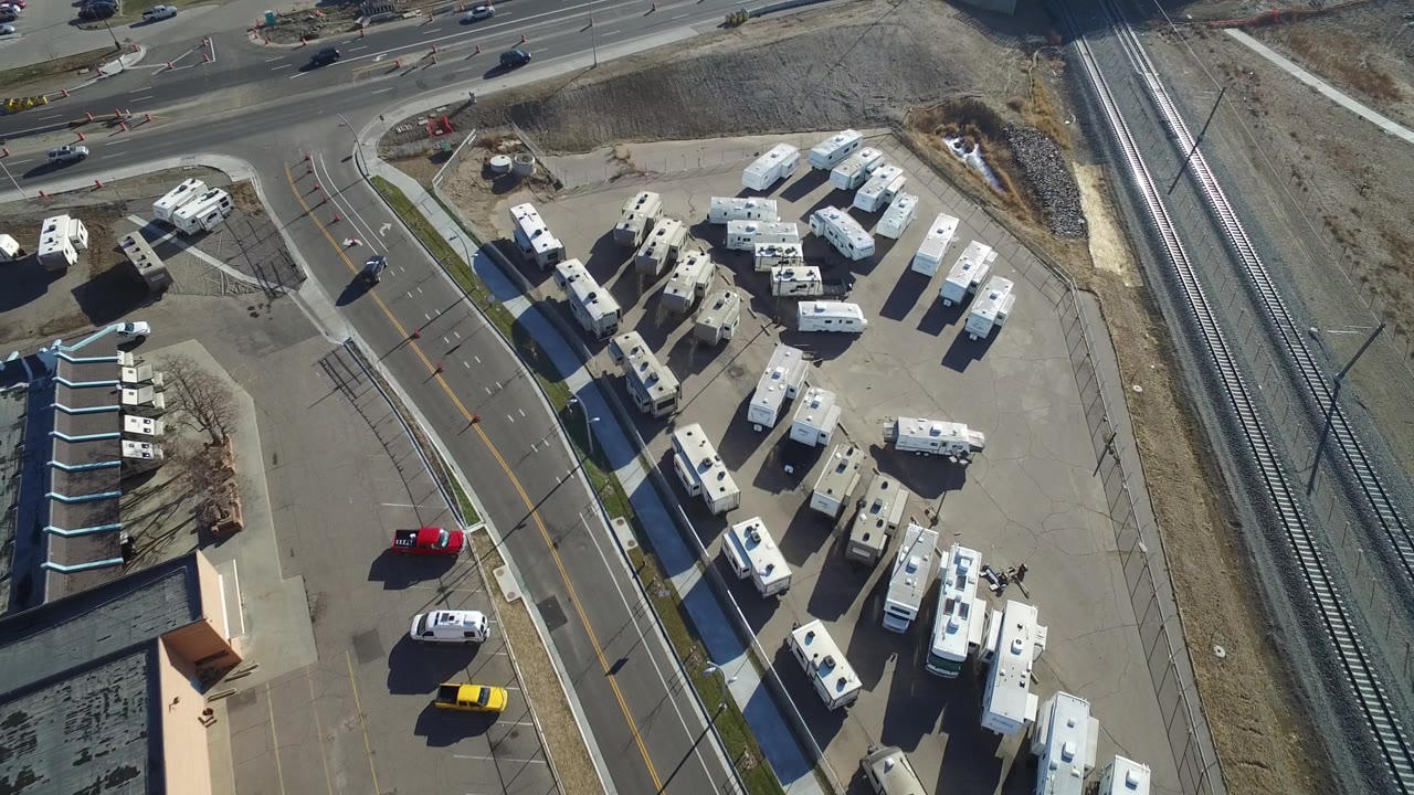 High overhead view in Hidden Lake, Colorado with rows of parked RVs, a red vehicle and a yellow vehicle on the road, rail tracks along the right side, and no visible people.