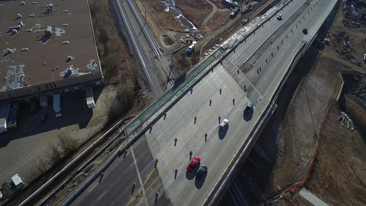High overhead view in Hidden Lake, Colorado showing an elevated road with traffic cones, a red car, rail tracks, a flat-roof building, dry ground, snow patches, and no visible people.