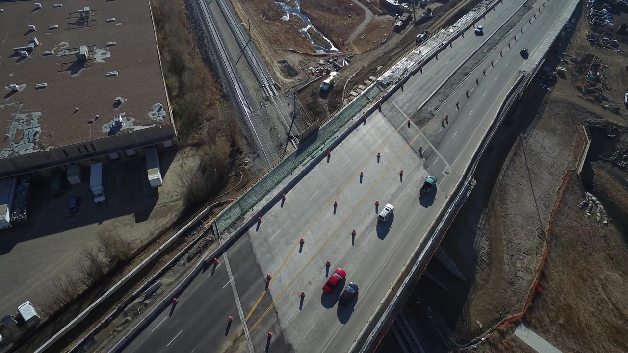 Aerial frame in Hidden Lake, Colorado with a road bridge running diagonally past rail tracks, scattered traffic cones, several vehicles, a brown-roof building, and no visible people.