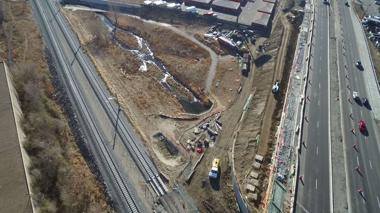 Aerial frame over Hidden Lake, Colorado showing highway lanes with cars, railroad tracks, dry vegetation, a drainage area, and industrial buildings, with no visible people.
