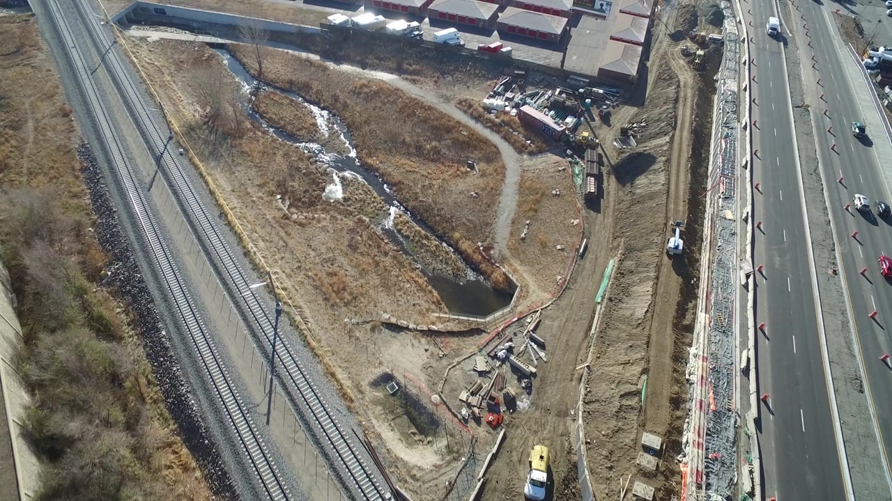 Aerial view over Hidden Lake, Colorado showing the highway shifted farther right in frame, with rail tracks, a curved dirt path, scrub vegetation, ponded water, and industrial yards, with no visible people.