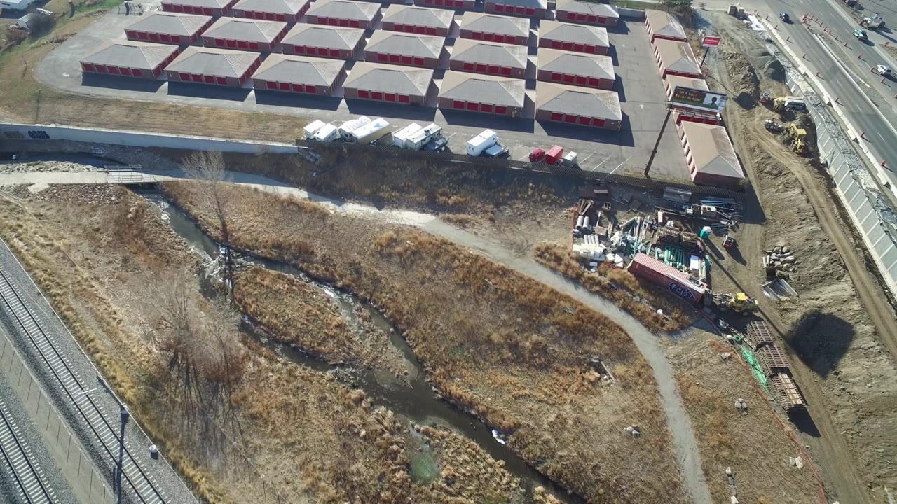Aerial view in Hidden Lake, Colorado with storage-unit rows across the top, a road with small vehicles, rail tracks at left, and scrubby drainage land below, with no clearly visible people.
