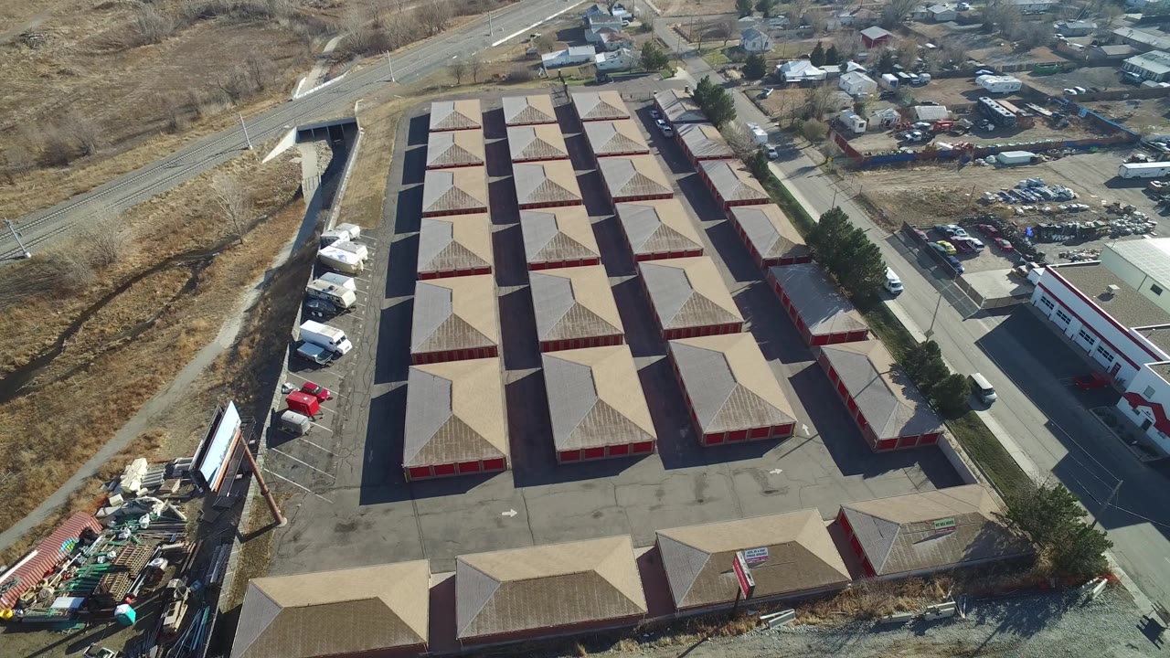 Opening aerial frame over an industrial building group in Hidden Lake, Colorado, with tan roofs, red walls, paved lanes, parked vehicles, dry open ground, and no visible people.