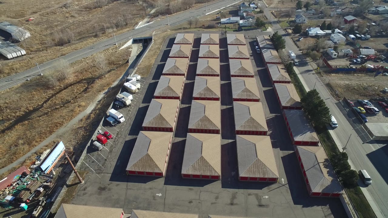 High aerial frame over a grid of tan-roofed buildings in Hidden Lake, Colorado, with parked vehicles along the left edge, nearby homes in the background, and no visible people.
