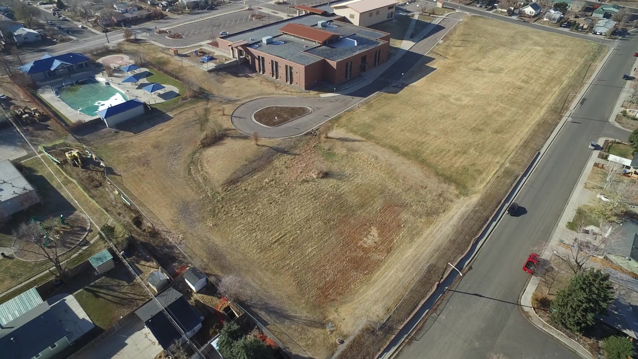 Aerial frame over Hidden Lake, Colorado showing a paved road, dry open grass, a brick building, nearby homes, trees, and no visible people.