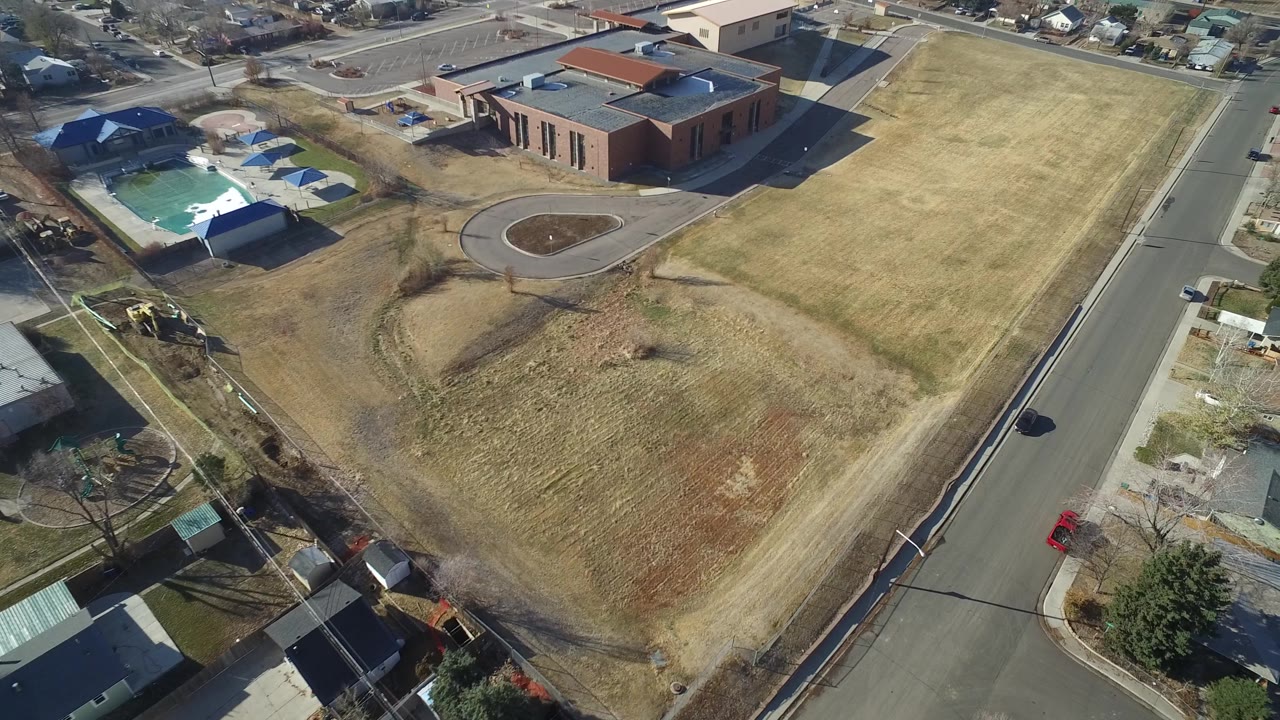 Aerial frame over Hidden Lake, Colorado with the brick building nearer the upper center, a long road at right, dry lawns, houses, and no visible people.