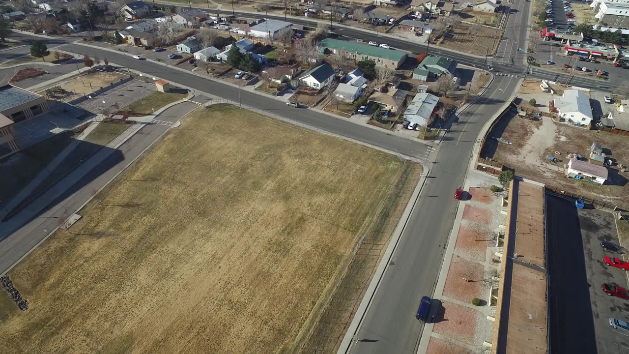 Opening aerial frame over Hidden Lake, Colorado showing a dry green field beside paved roads, low buildings, small homes, parked vehicles, and no visible people.