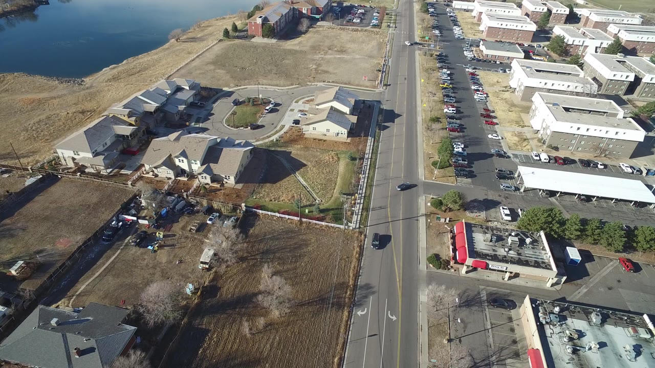 Opening aerial frame over Hidden Lake, Colorado with homes in the lower left, a straight road through the center, industrial buildings on the right, lake water at the upper left, and no visible people.