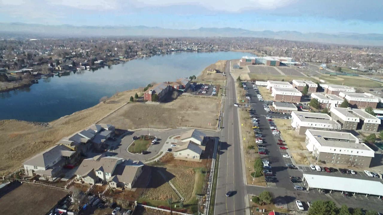 Wide aerial view of a lake, road, buildings, suburbs, and distant mountains.