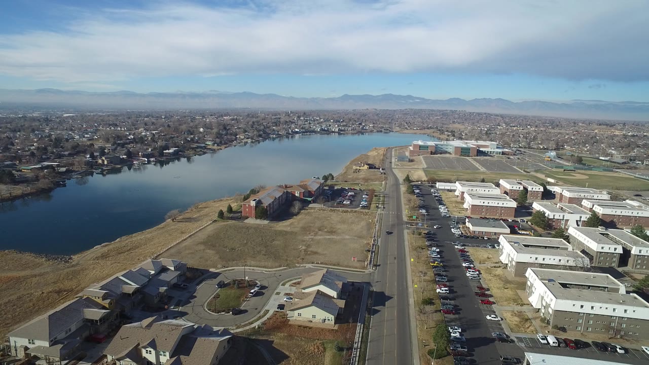 Opening aerial frame over Hidden Lake, Colorado showing the lake on the left, a straight road through the center, low buildings and parked cars on the right, distant neighborhoods and mountains, and no visible people.