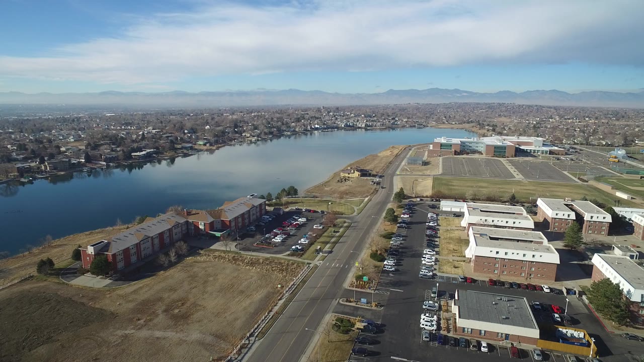 Aerial frame from Hidden Lake, Colorado showing apartment buildings and parking lots in the foreground, a road leading toward the lake, distant neighborhoods, mountains, thin clouds, and no visible people.