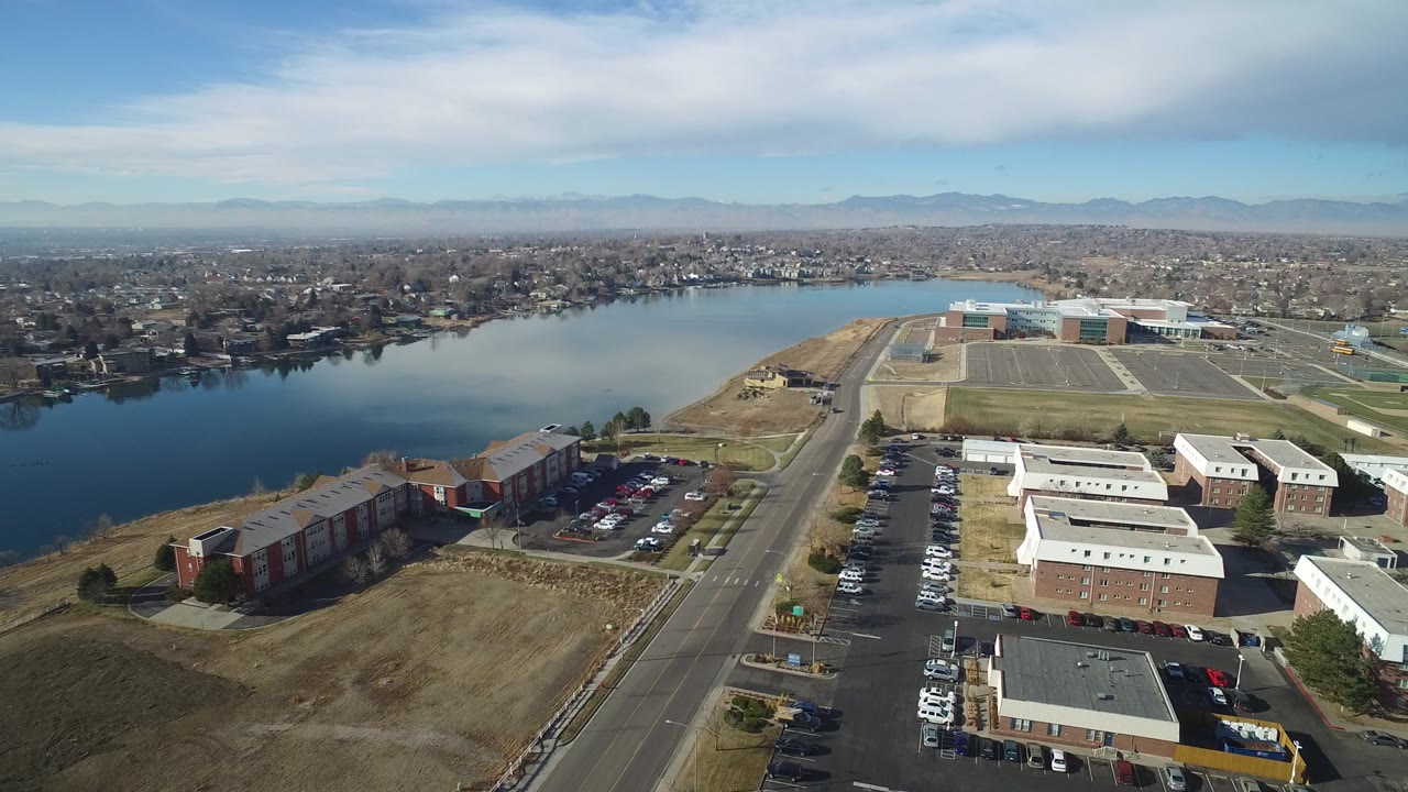 Aerial view in Hidden Lake, Colorado with the lake across the middle distance, apartment buildings and parked cars below, a main road near center, distant urban housing, mountains, and no visible people.