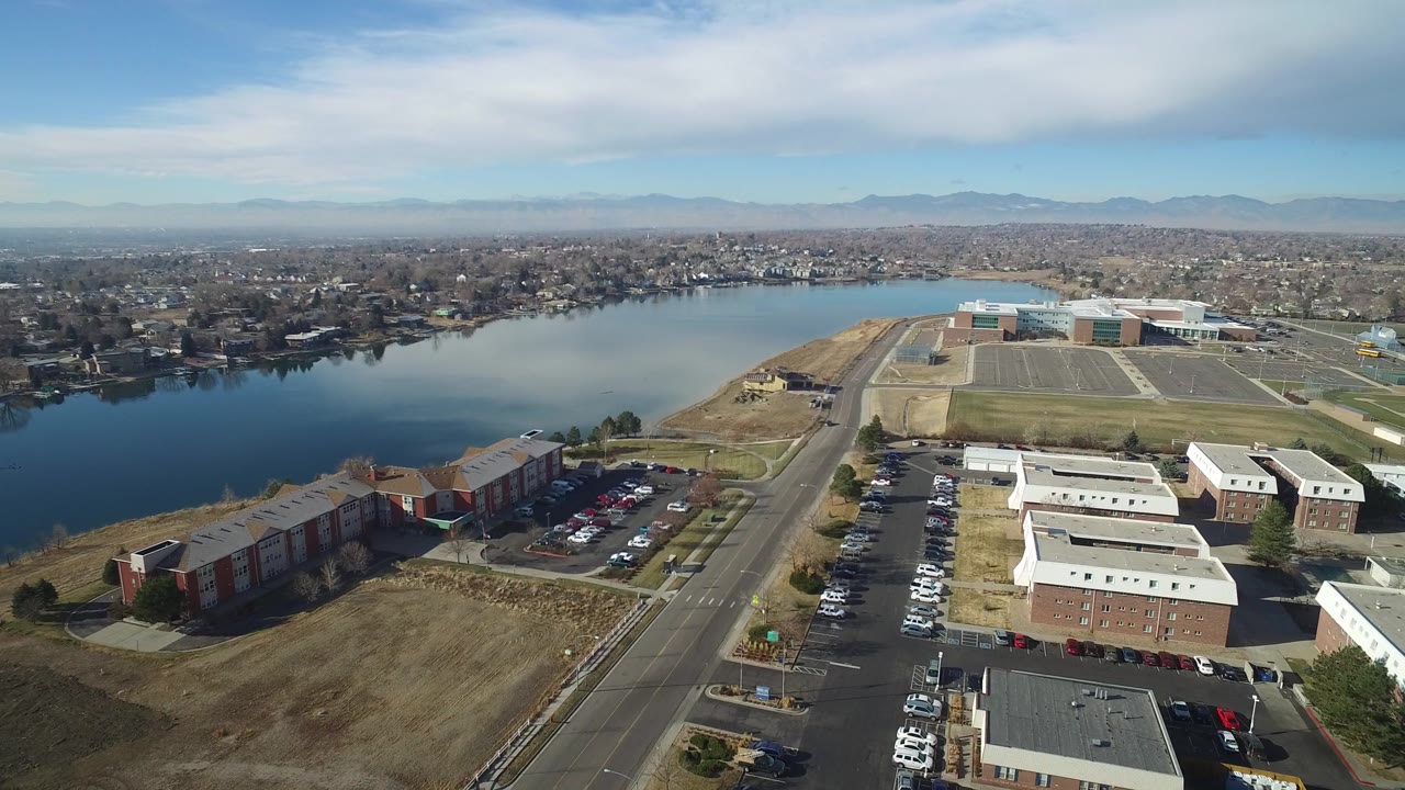 Wide aerial of a lake, apartment complex, parking lots, road, and distant mountains.