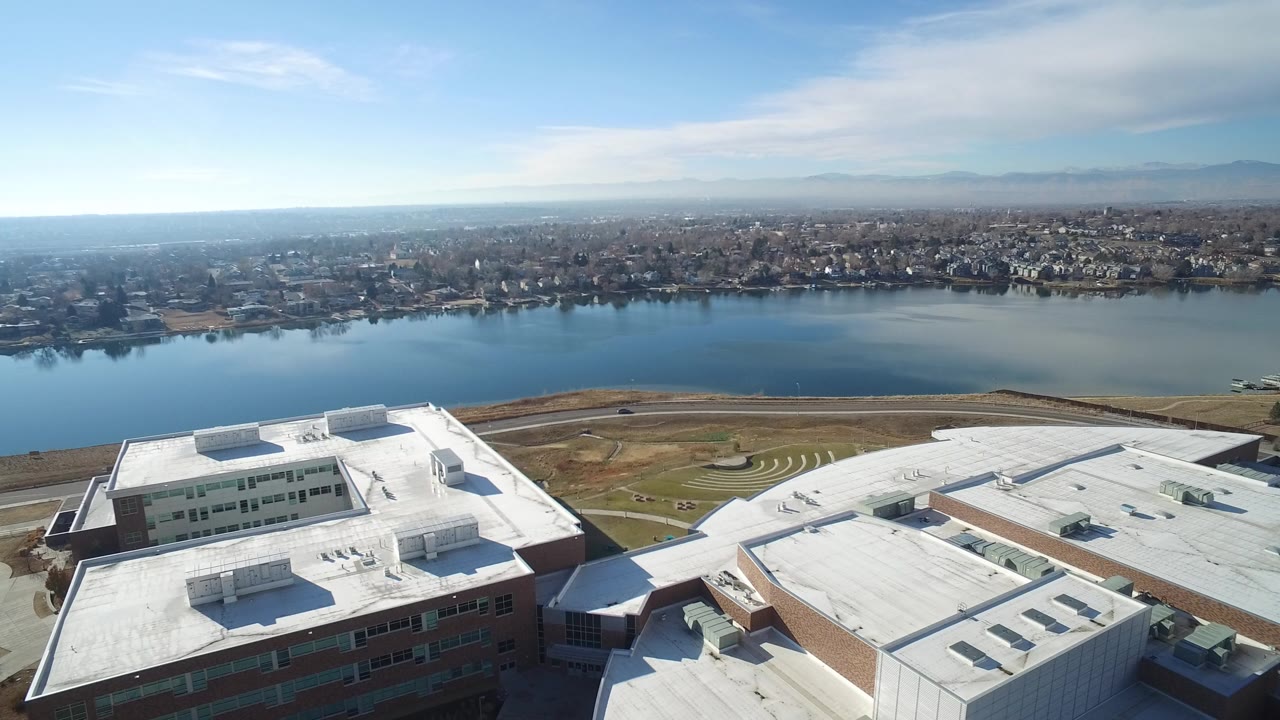 Aerial frame over Hidden Lake, Colorado showing white flat rooftops in the foreground, calm water beyond, houses across the shoreline, distant mountains, and no visible people.