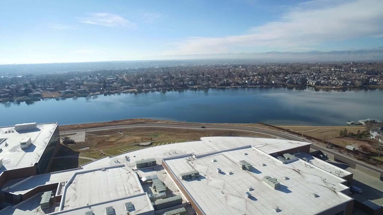 Aerial view over white rooftops toward a blue lake and distant neighborhood.