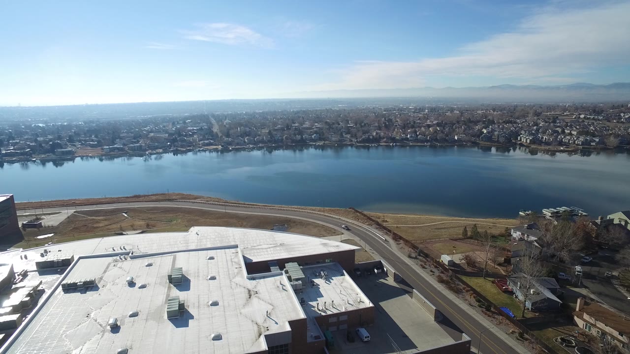 Opening aerial frame over Hidden Lake, Colorado with a bright white commercial-style roof at lower left, a curved road, blue lake water, distant homes, hazy mountains, and no visible people.