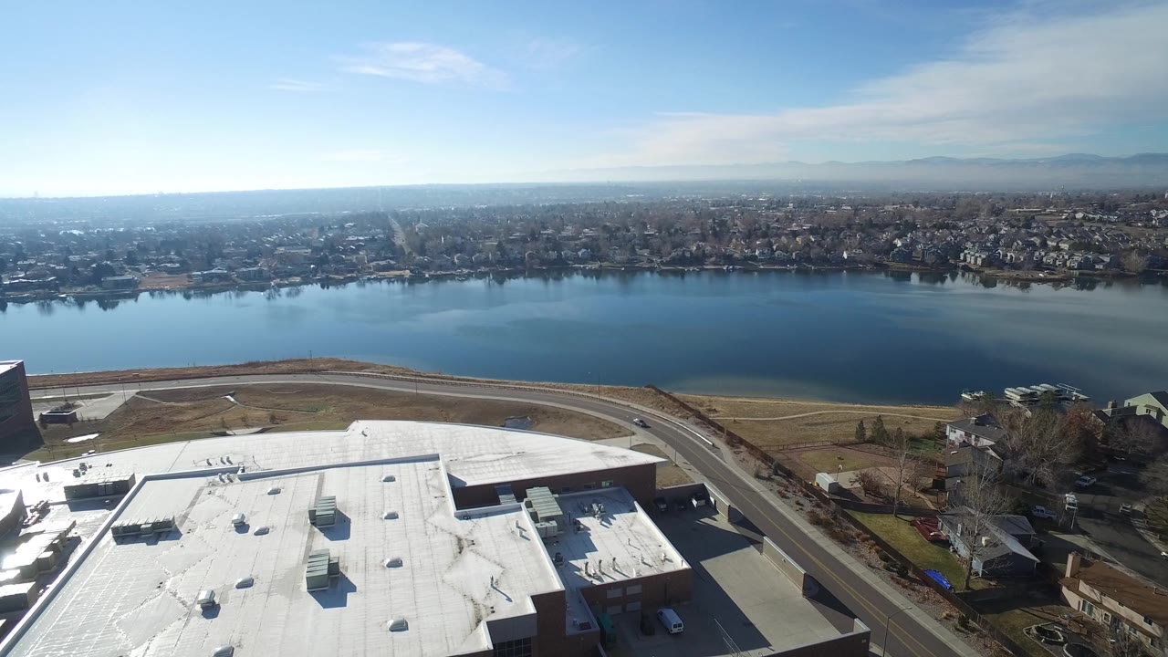 Aerial frame in Hidden Lake, Colorado looking across the lake from a foreground white roof and shoreline road toward residential blocks on the far bank, with no visible people.