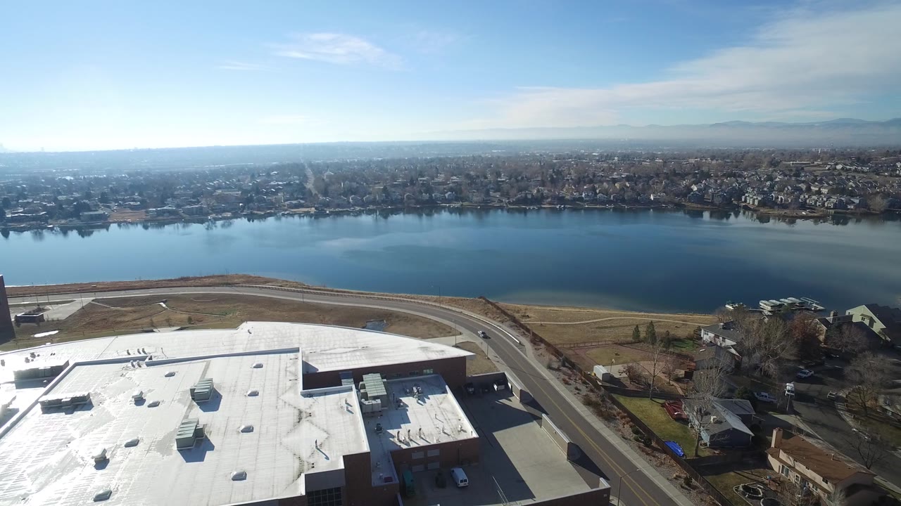 Wide aerial over a blue lake, road, white rooftop, houses, and pale mountains.