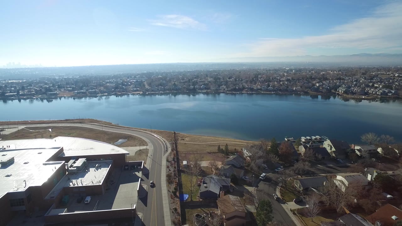 Aerial view in Hidden Lake, Colorado, with the lake centered, a pale shoreline path and road below, dense residential blocks across the water, and no visible people.