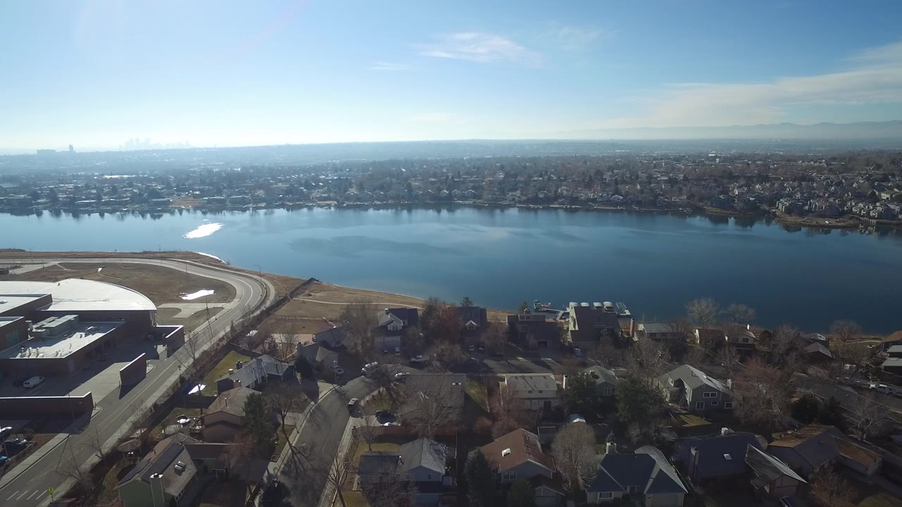 Wide aerial view of a lake, neighborhood rooftops, roads, and distant shoreline.