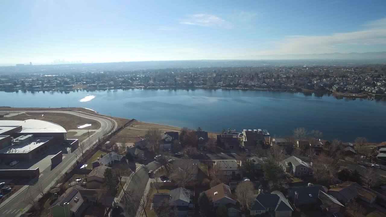 Aerial view over Hidden Lake, Colorado, with a curved road and large flat-roofed building at left, houses below, reflective water across the center, and no visible people.