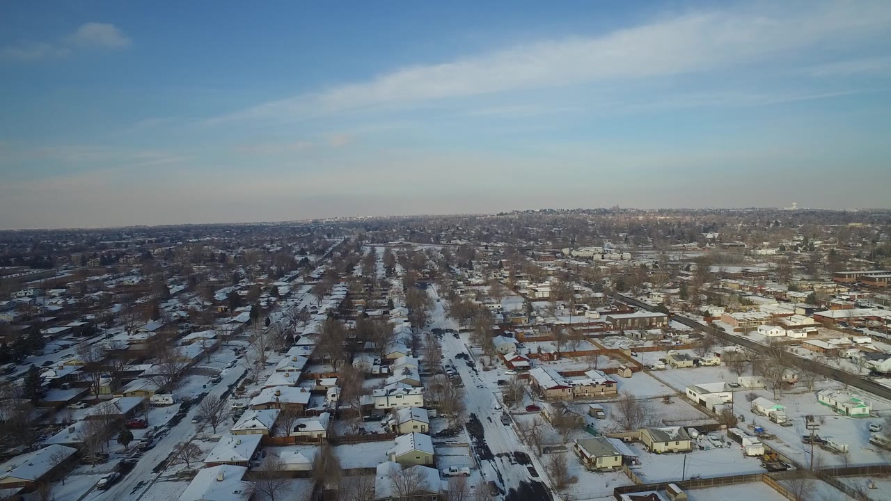 Drone perspective over snowy homes with commercial buildings in the distance.