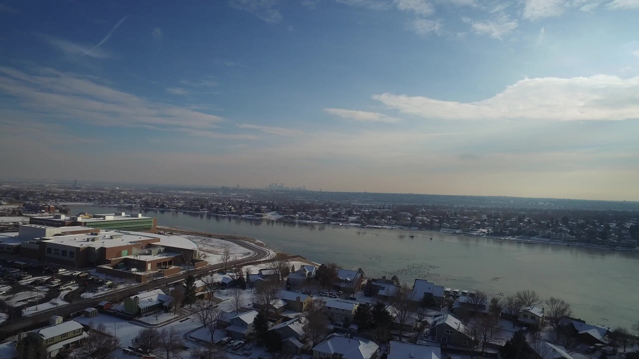 Opening frame of Hidden Lake, Colorado showing snow-covered rooftops in the foreground, a low shoreline building, broad water, distant houses, and no visible people.