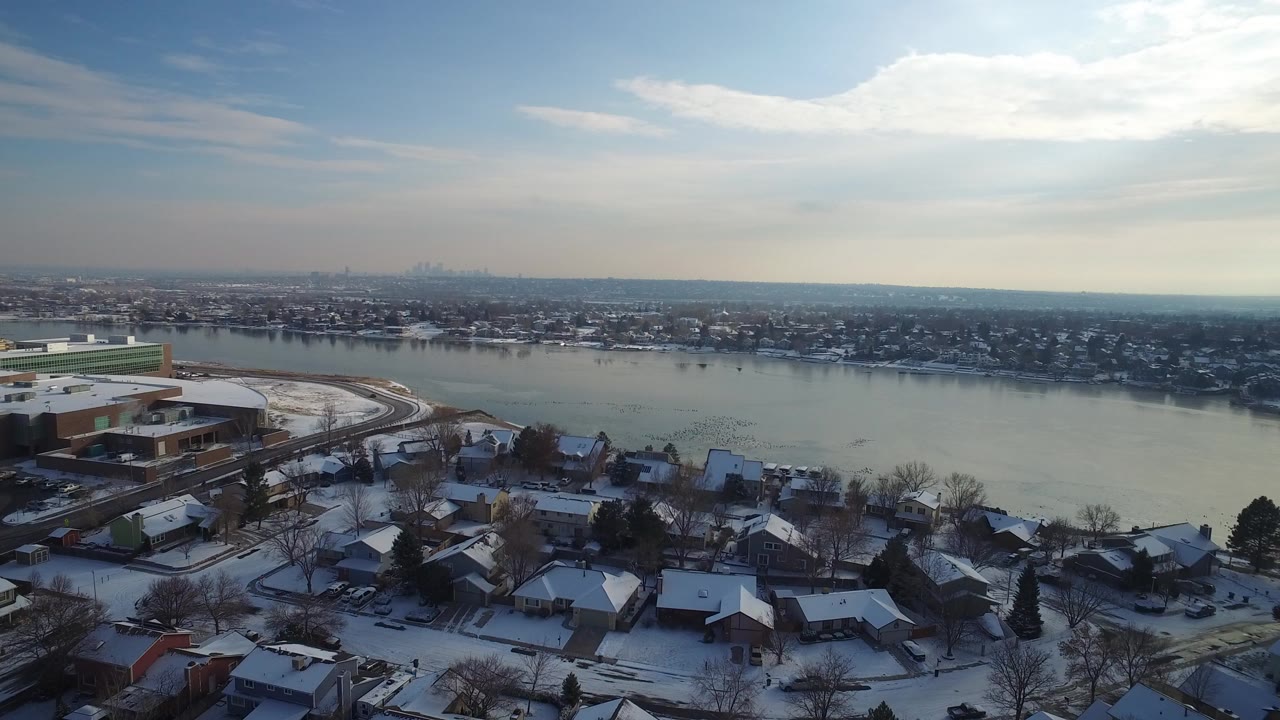 Opening aerial frame over Hidden Lake, Colorado, showing snow-covered rooftops in the foreground, a broad waterway in the middle distance, a hazy skyline on the horizon, and no visible people.