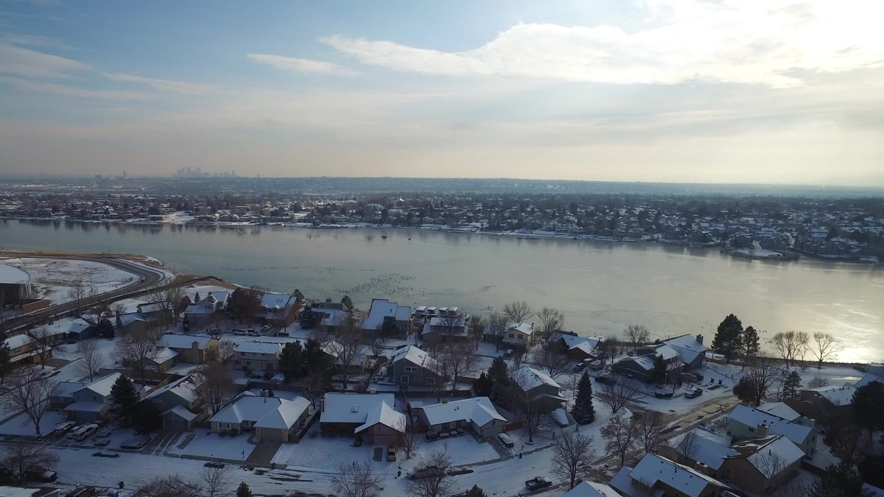 Aerial view of Hidden Lake, Colorado with snowy rooftops in the foreground, a reflective lake across the center, distant neighborhoods on the horizon, and no visible people.