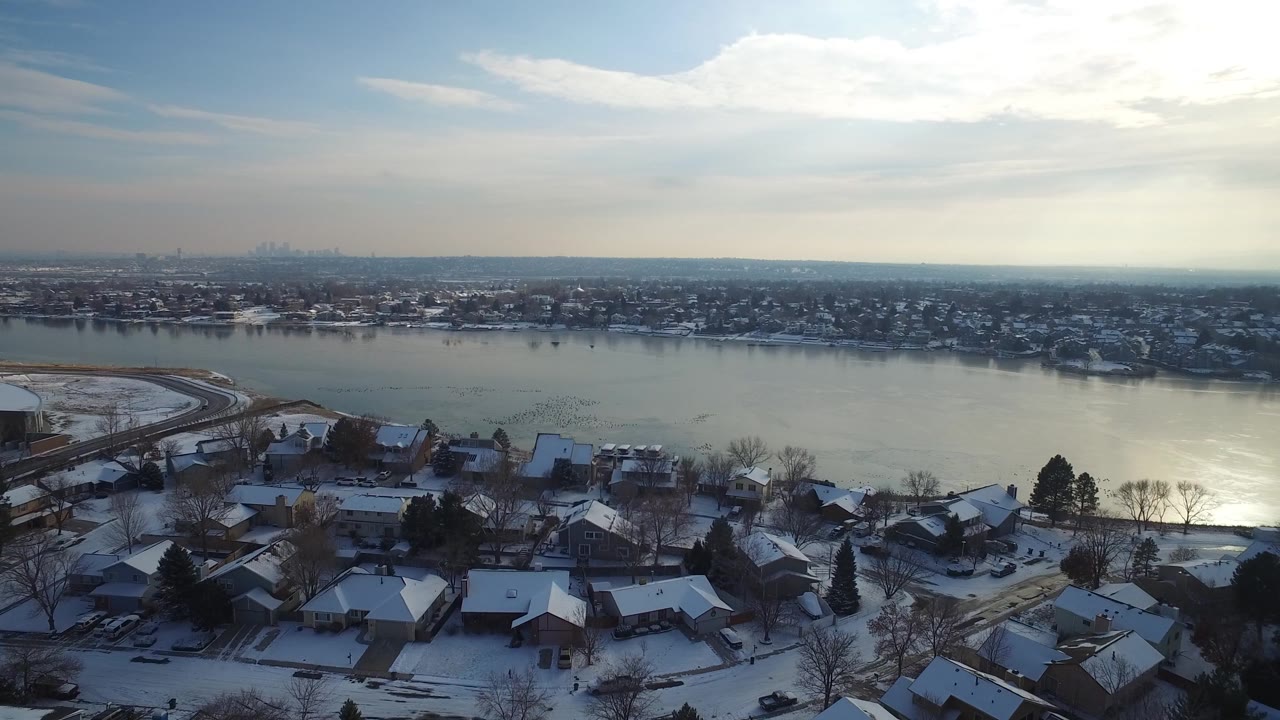 Hidden Lake, Colorado appears from above with clustered snow-covered homes below, a curved shoreline road at left, bright winter light on the lake, and no visible people.