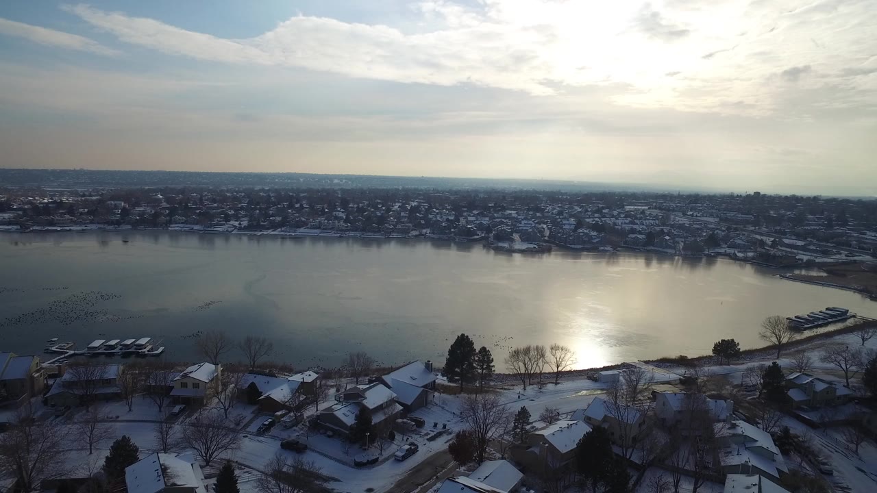 Snowy homes beside a lake with sun glare on the water.