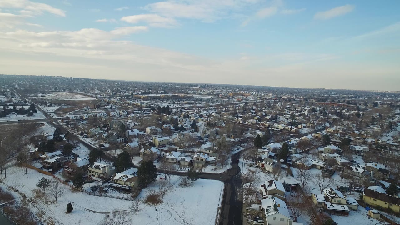 Snowy neighborhood aerial with homes, trees, streets, and distant low buildings.