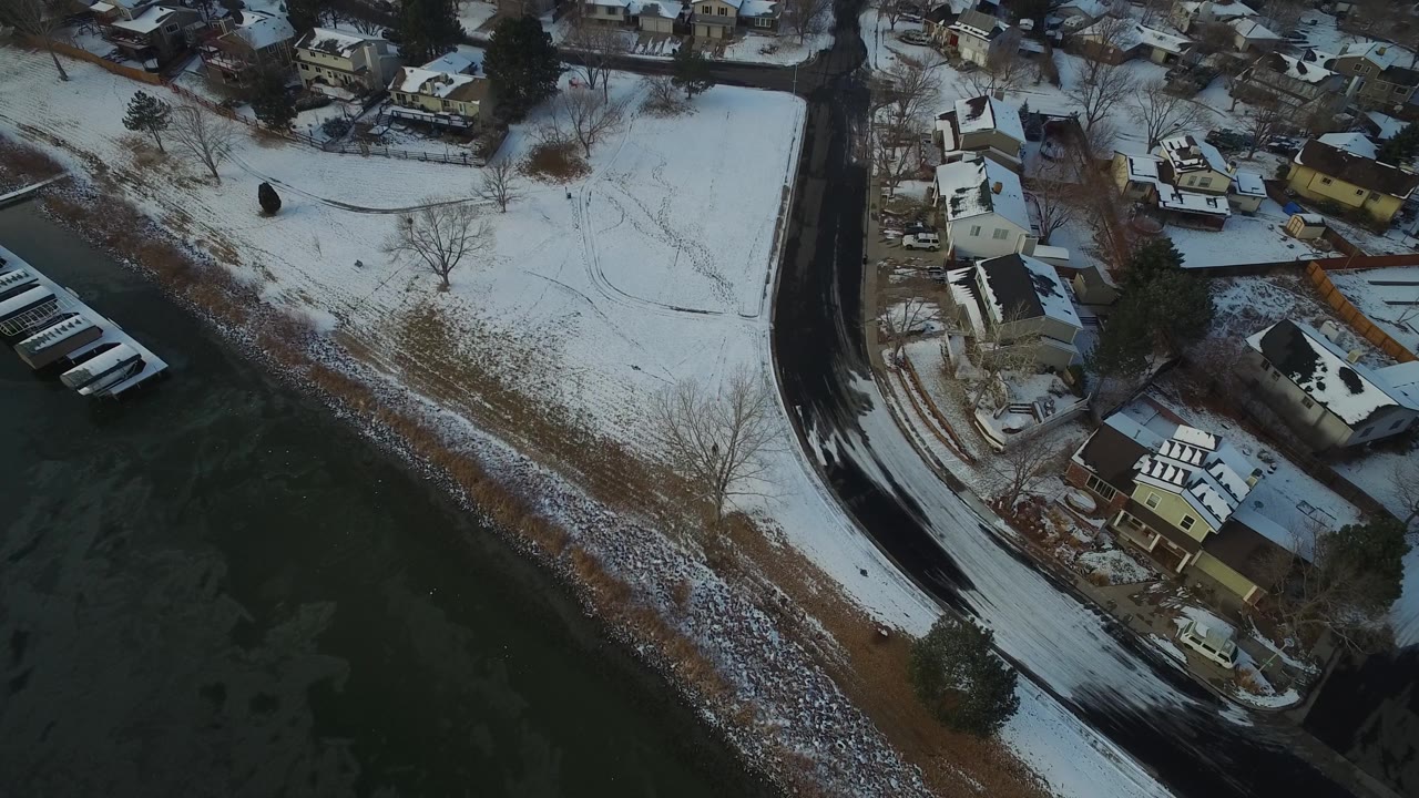 Aerial view in Hidden Lake, Colorado, with the snowy open ground and road shifted across frame while lake water and residential houses remain visible, with no visible people.