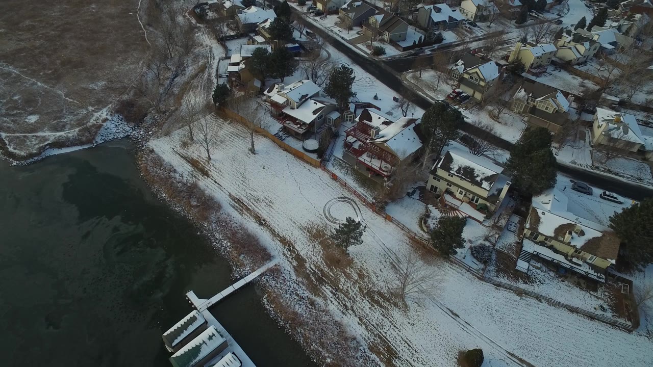 Top-down aerial of snow-dusted homes, shoreline, dock, and open terrain.