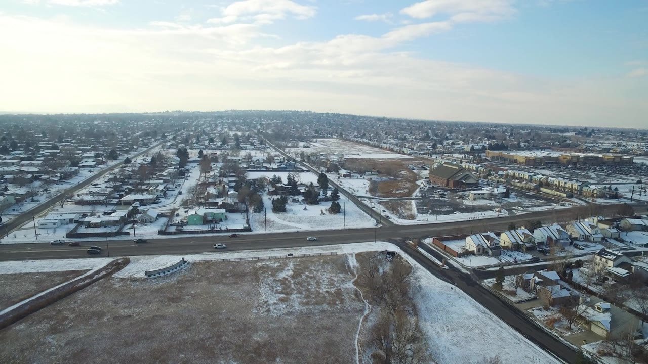 Aerial view of snowy roads, houses, open lots, and a distant neighborhood under cloudy sky.