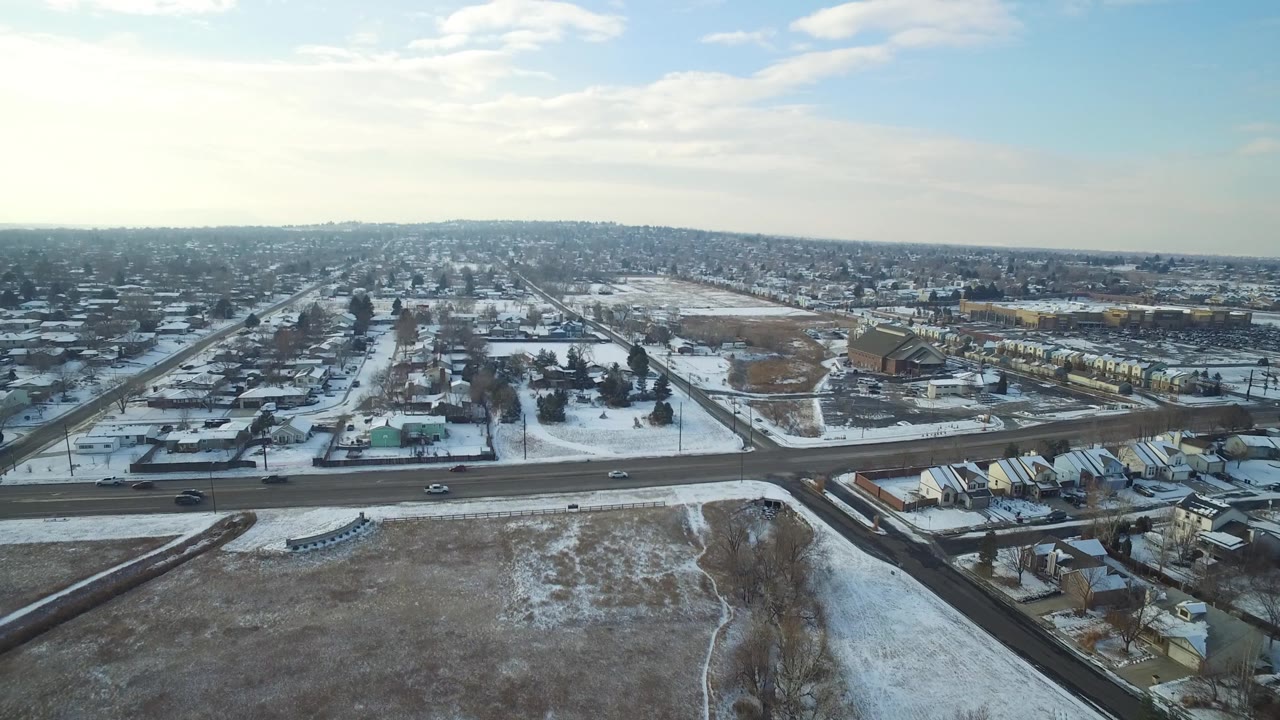 Aerial view in Hidden Lake, Colorado, with a broad road crossing the lower frame, small cars in traffic, rows of houses, an open snowy tract, distant buildings, and no visible people.
